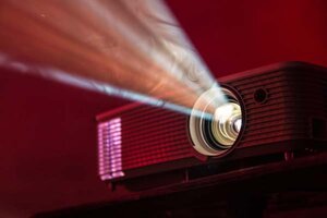 LED-Projektor auf dem Tisch eingeschaltet; Bild: Alex Litvin auf Unsplash Bild von einem Beamer, der in der Dunkelheit stahlt.