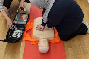 Mann in schwarzem Pullover und weißer Hose sitzt auf braunem Holztisch; Bild: Martin Splitt auf Unsplash Frau übt Herzdurckmassage an einer Puppe.